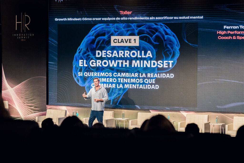 Fotografía de Ferran Tort en el escenario de una conferencia, presentando sobre desarrollo del growth mindset con una gran pantalla de fondo que muestra un cerebro en azul y mensajes motivacionales.