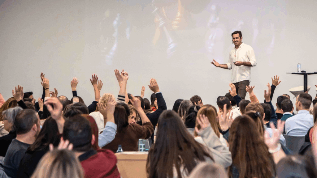 Fotografía de Ferran Tort en un escenario interactuando con una audiencia entusiasta, donde numerosos asistentes levantan la mano durante su conferencia.