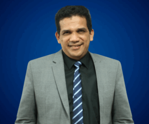 Fotografía de Jaime Montenegro, con fondo azul sólido, vistiendo traje gris, camisa negra y corbata a rayas, mostrando una expresión amable y profesional.