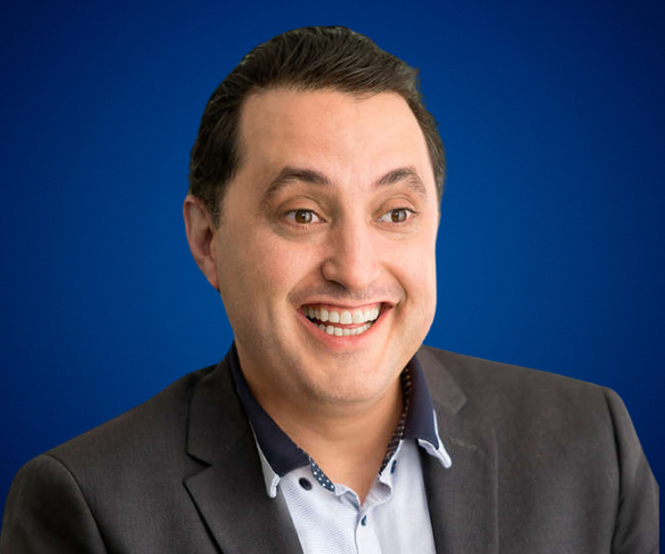 Retrato profesional de un hombre sonriente con traje oscuro y camisa blanca, sobre un fondo azul.