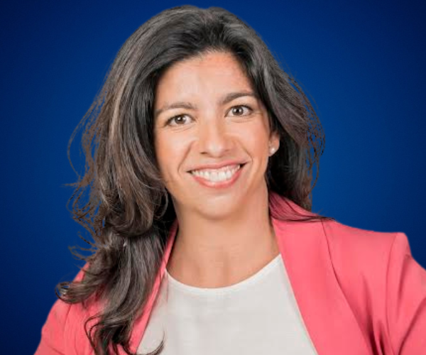 Marta Freire sonriendo, vistiendo un blazer rosa y una blusa blanca, sobre un fondo azul.