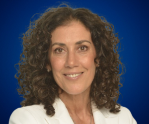 Paula Dinaro sonriendo, experta en bienestar y liderazgo consciente, con fondo azul.