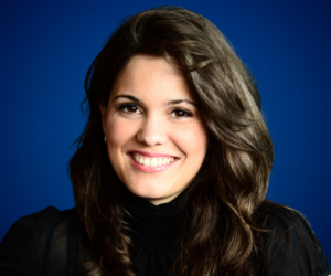 Retrato de Ana María De la Guardia, con cabello ondulado y vestimenta negra, sonriendo sobre un fondo azul.