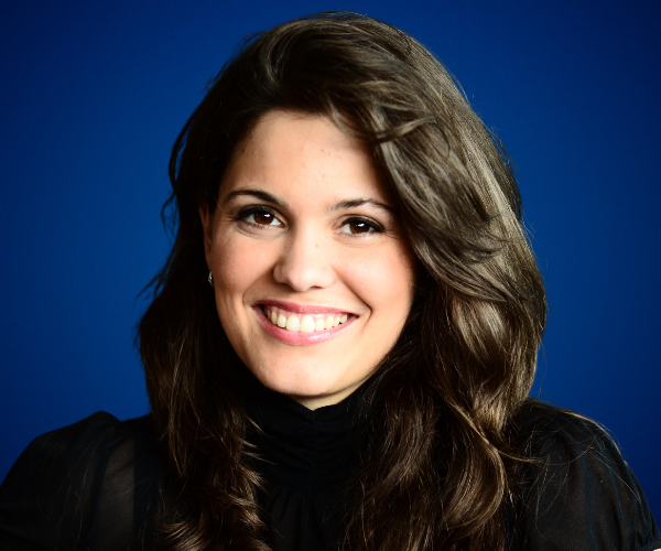 Retrato de Ana María De la Guardia, con cabello ondulado y vestimenta negra, sonriendo sobre un fondo azul.