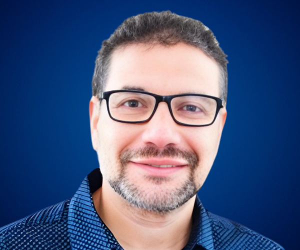 Retrato profesional de Luis Manuel Ramírez con fondo azul, vistiendo una camisa azul con patrón discreto y gafas negras, transmitiendo confianza y profesionalismo.