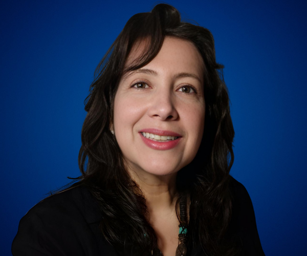 Retrato de María Carolina Villalobos, conferencista y coach, sonriendo con cabello oscuro sobre un fondo azul, vistiendo atuendo profesional.