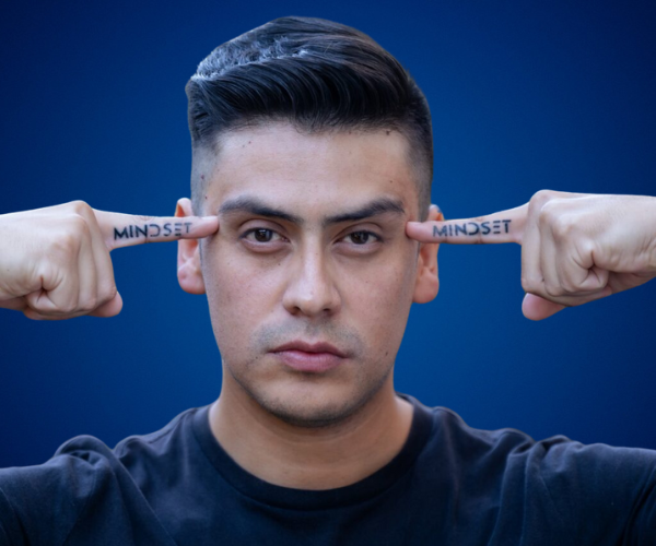 Retrato de Manuel Vásquez Arce apuntando con los dedos a sus sienes, donde tiene tatuada la palabra "MINDSET", con fondo azul y expresión decidida.