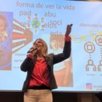 Vivianne Verschae ofreciendo una charla con micrófono en mano frente a una presentación proyectada sobre creencias y roles sociales.