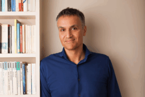 Carl Honoré de pie junto a una estantería de libros, luciendo una camisa azul, en un ambiente cálido e intelectual.