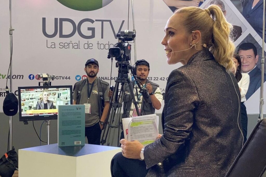 Fotografía de Adriana Lozano en una entrevista televisiva, usando micrófono de diadema y sosteniendo documentos, frente a cámaras y equipo técnico de UDGTV.