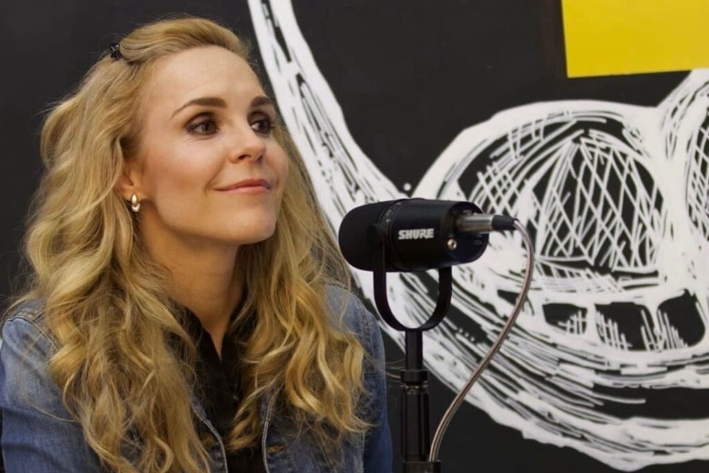 Fotografía de Adriana Lozano durante una entrevista o conferencia, sonriendo con serenidad frente a un micrófono profesional, sobre un fondo artístico en blanco y negro.
