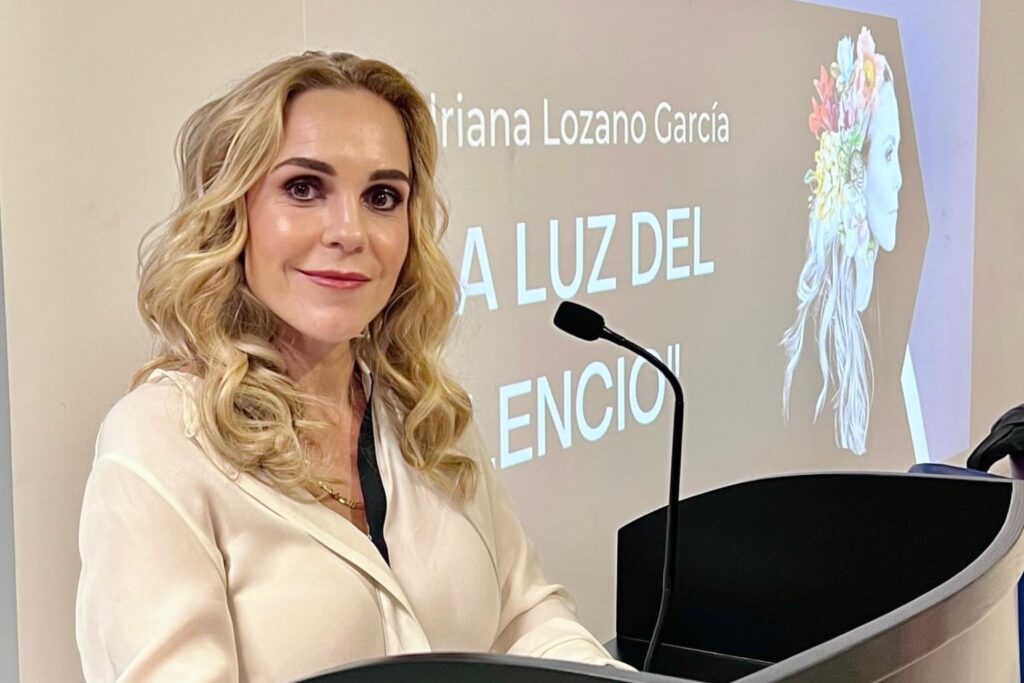 Fotografía de Adriana Lozano durante una conferencia, de pie tras un podio, con fondo de proyección titulado "La luz del silencio".