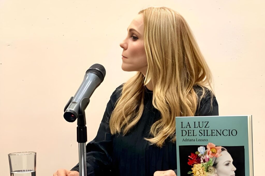 Fotografía de Adriana Lozano durante una presentación, sentada junto a un micrófono y su libro “La luz del silencio”.