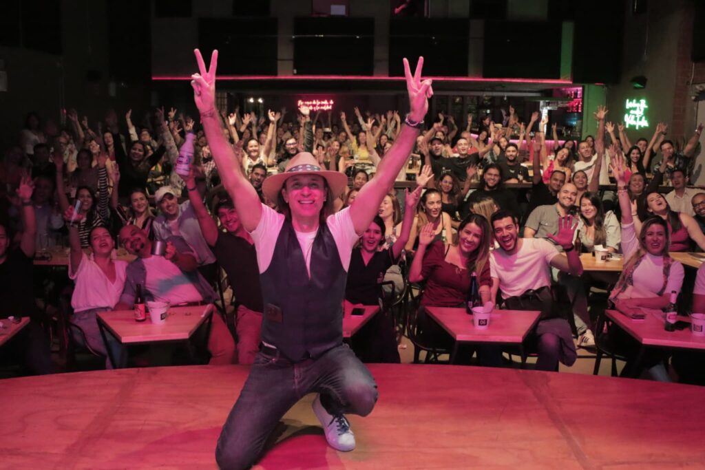 Fotografía de Ezequiel López en el escenario de un evento en vivo, sonriendo con entusiasmo y levantando ambas manos en señal de victoria, rodeado de un público alegre que celebra con él.