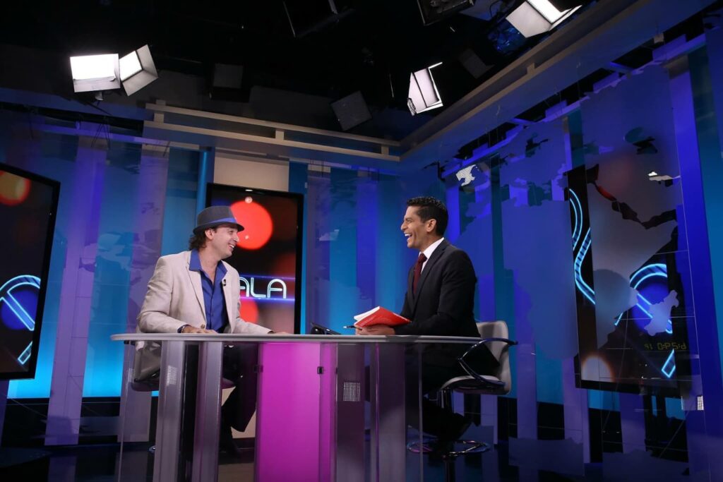 Fotografía de Ezequiel López en una entrevista televisiva dentro de un estudio moderno, conversando con un presentador mientras sostiene un libro rojo.