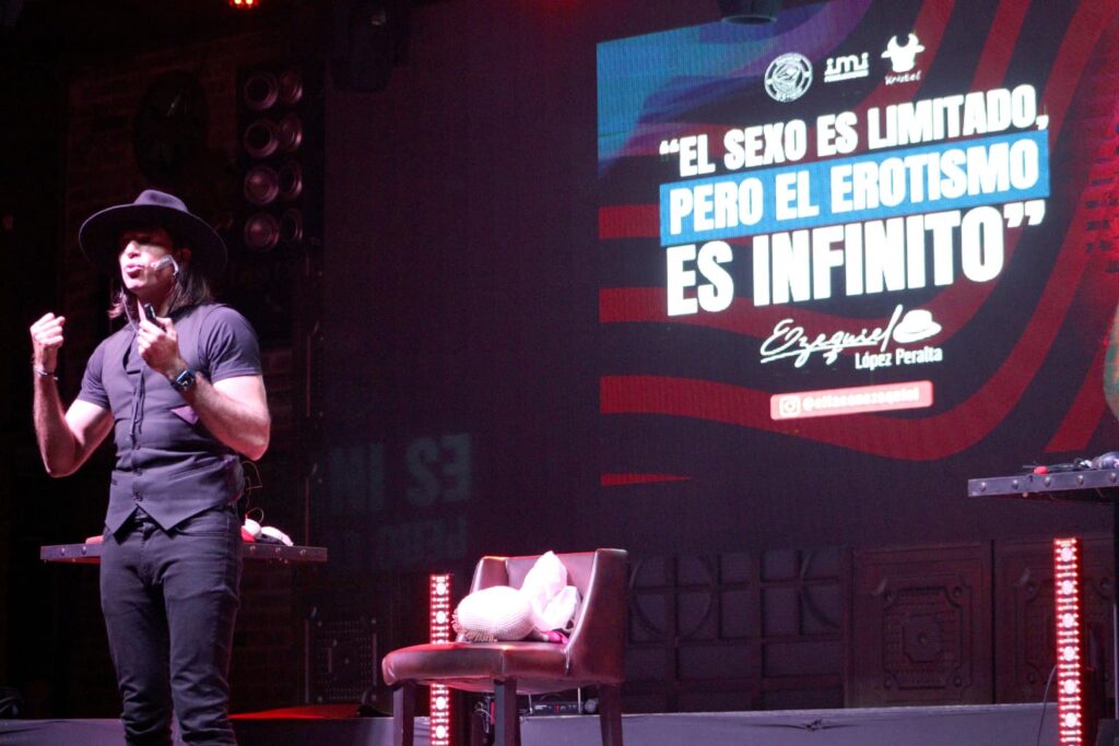 Fotografía de Ezequiel López Peralta en el escenario durante una conferencia, con sombrero negro y micrófono, acompañado de una proyección con la frase: “El sexo es limitado, pero el erotismo es infinito”.
