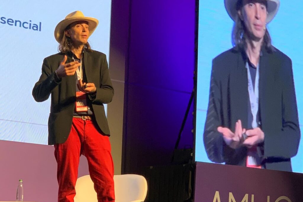 Fotografía de Ezequiel López Peralta dando una conferencia, vestido con sombrero blanco, saco oscuro y pantalón rojo, mientras expone frente a un público con una pantalla gigante proyectando su imagen.