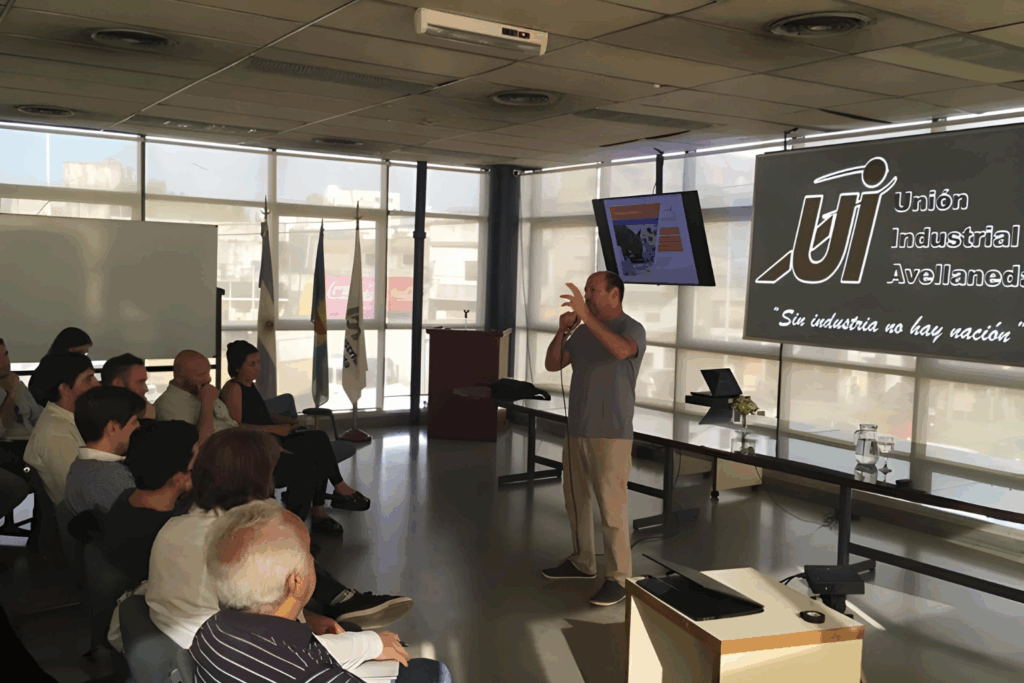 Fotografía de Pablo Nachtigall dando una charla en la Unión Industrial de Avellaneda, frente a un público atento. La imagen muestra un salón moderno con luz natural, banderas institucionales y una pantalla con presentación proyectada.