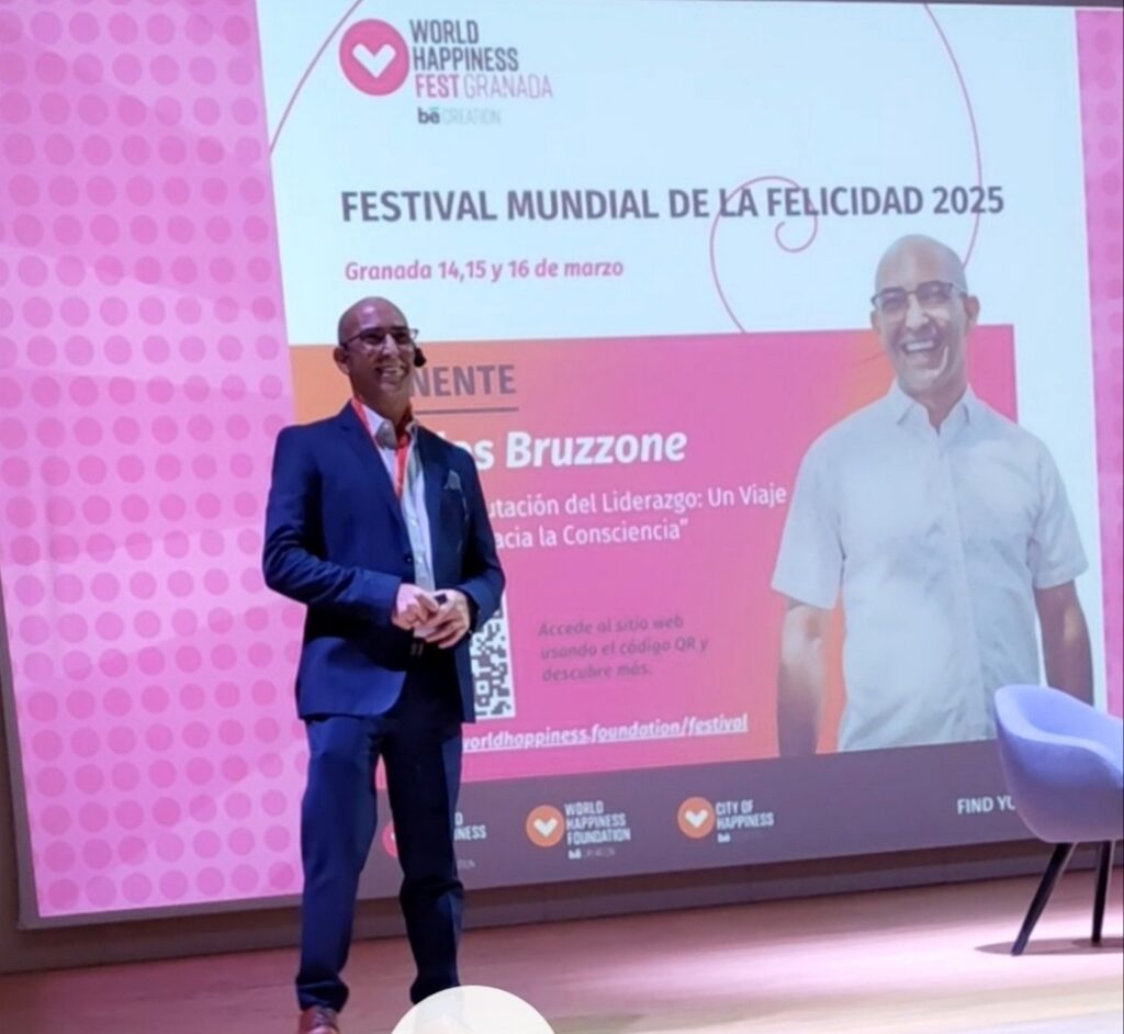 Fotografía de Carlos Bruzzone en el escenario del Festival Mundial de la Felicidad 2025 en Granada, vestido con traje azul y gafas, frente a una pantalla que anuncia su conferencia.