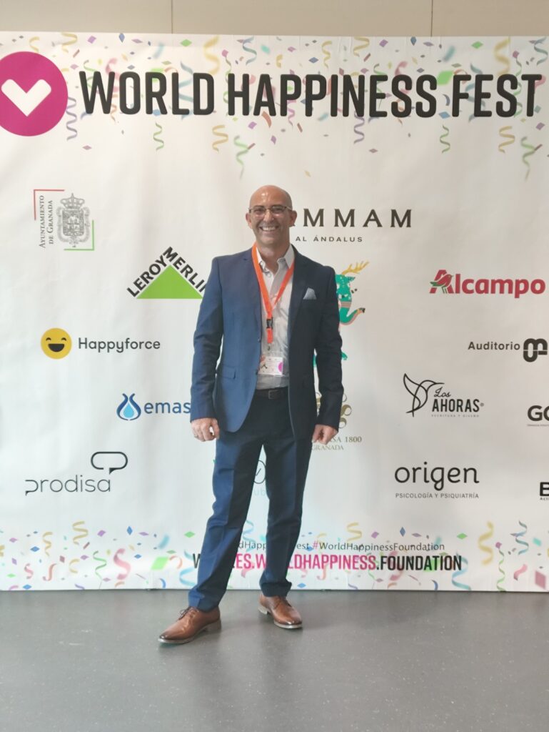 Fotografía de Carlos Bruzzone en el World Happiness Fest, posando con traje azul y gafas frente a un fondo promocional con logos de patrocinadores y decoraciones festivas.