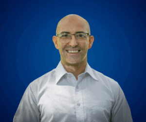 Fotografía de Carlos Bruzzone con gafas y camisa blanca, sonriendo frente a un fondo azul liso con iluminación suave.