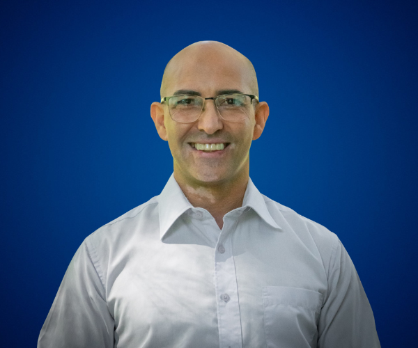 Fotografía de Carlos Bruzzone con gafas y camisa blanca, sonriendo frente a un fondo azul liso con iluminación suave.