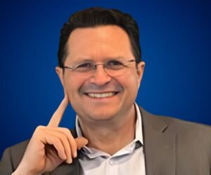 Fotografía de César Camacho, sonriendo con actitud cercana y profesional, vistiendo saco gris y camisa blanca sobre fondo azul sólido.