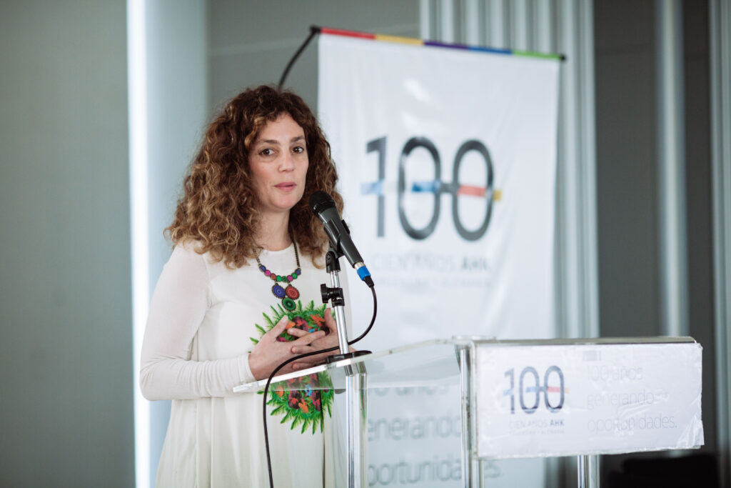 Débora Slotnisky hablando en un evento institucional de "100 Años AHK", de pie frente a un micrófono y podio transparente, con fondo corporativo blanco.