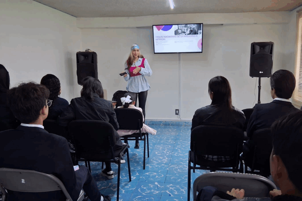 Fotografía de Brenda Caballero impartiendo una charla sobre el nacimiento de la inteligencia artificial, frente a un grupo de asistentes en un salón con pantalla y equipo de sonido.