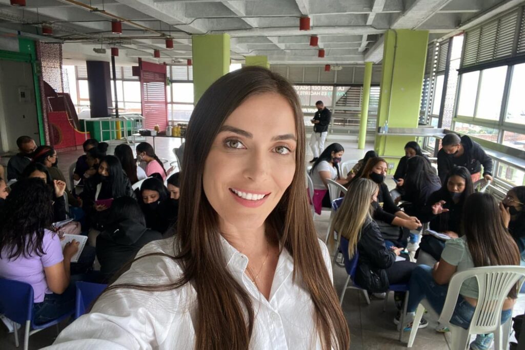 : Fotografía de Catalina Serna en primer plano, sonriendo mientras al fondo se observa un grupo de personas participando en una actividad formativa en un salón amplio y luminoso.