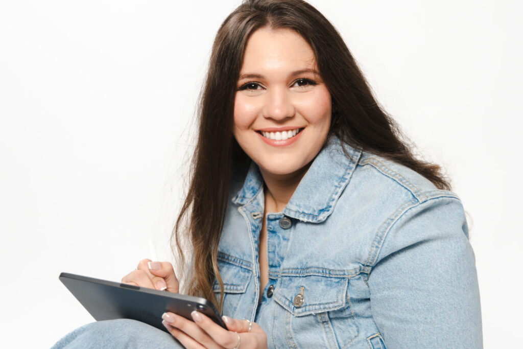 Fotografía de Danel Nouel con chaqueta de mezclilla, sonriendo mientras sostiene una tablet y un lápiz digital, sobre un fondo blanco.