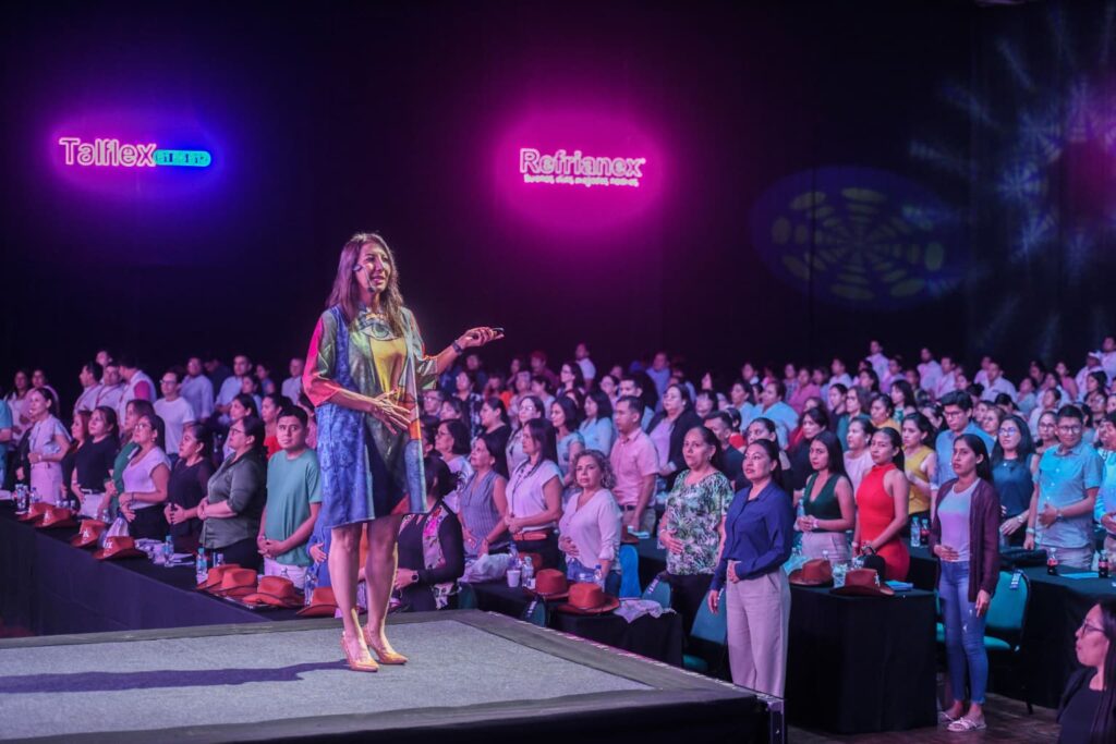Fotografía de María Antonieta Alcalá en el escenario durante una conferencia, vestida con un atuendo colorido y rodeada de un público de pie, bajo luces púrpuras.