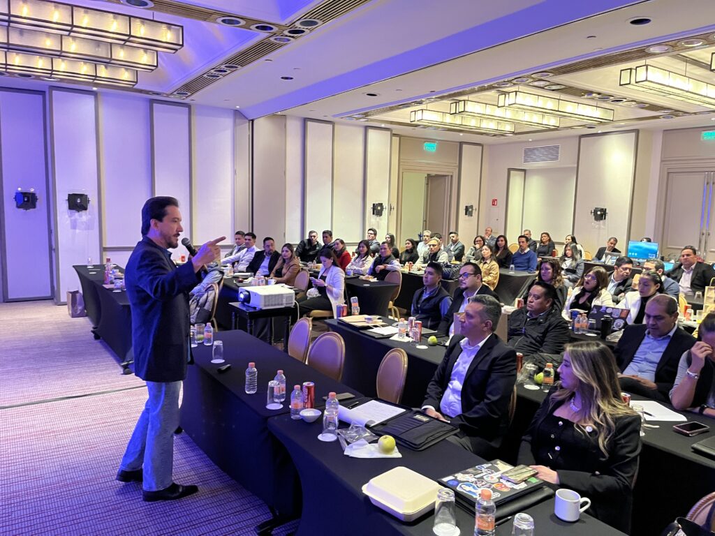 Ness Carballo impartiendo una conferencia en un salón de eventos, dirigiéndose a una audiencia atenta en un entorno profesional y corporativo.