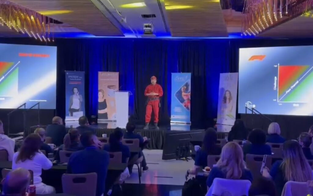Ness Carballo en un escenario de conferencia, vestido completamente de rojo, frente a dos pantallas con gráficos y banners promocionales, dirigiéndose a una audiencia atenta en un salón iluminado.