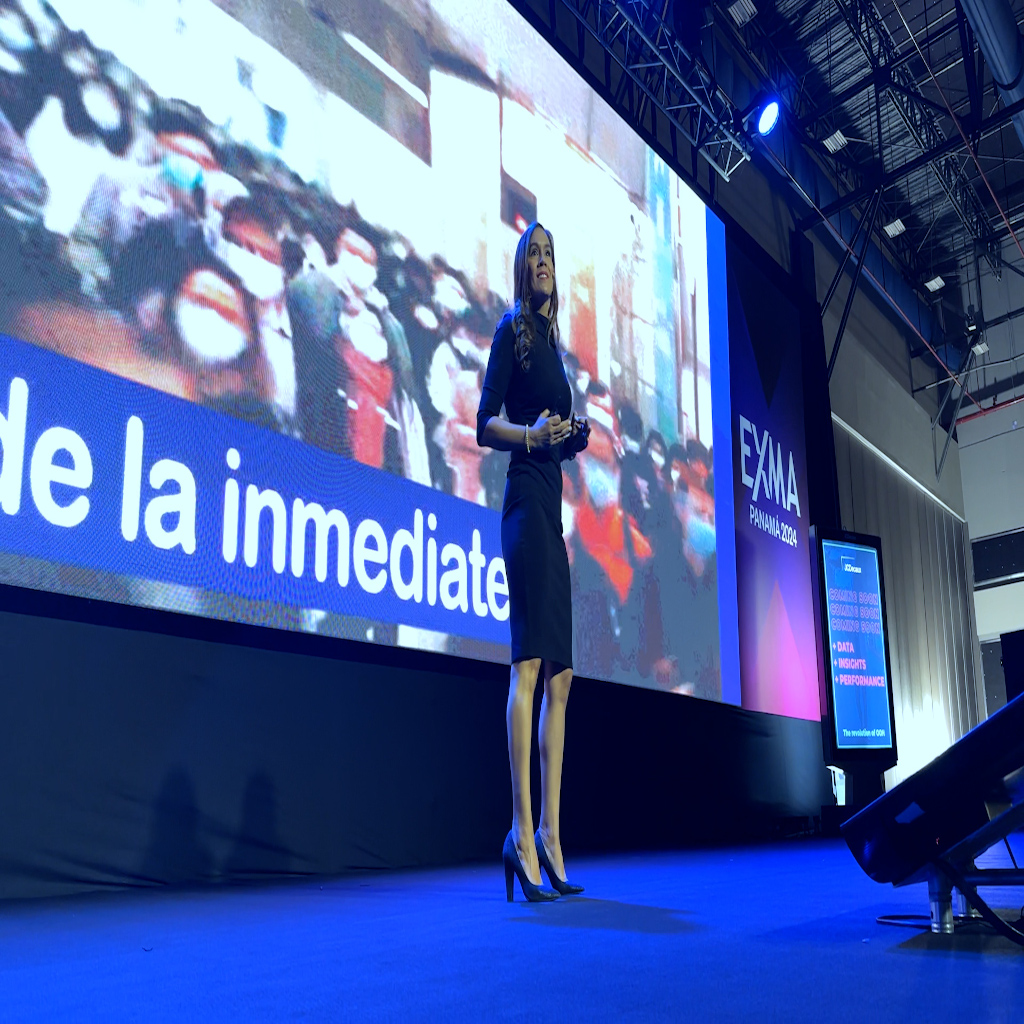 Fotografía de Nadia Vado en el escenario de EXMA Panamá 2024, vestida profesionalmente con traje oscuro, frente a una pantalla gigante con gráficos y textos, bajo iluminación azul y ambiente de gran evento.