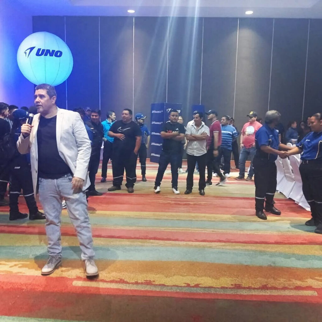 Fotografía de Rodrigo Quintanilla hablando con micrófono durante un evento corporativo de la marca UNO, con numerosos asistentes en un salón iluminado con luces azules.