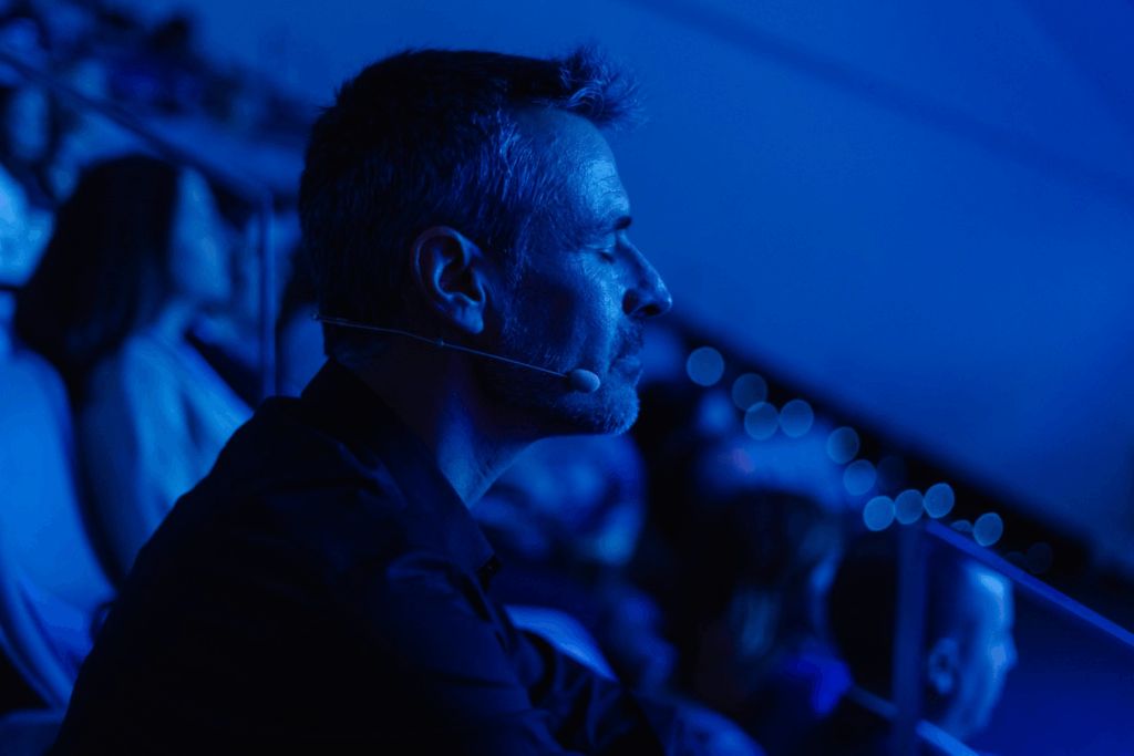 Fotografía de Antoni Fernández i Brugués en perfil, sentado entre el público durante un evento, iluminado por una intensa luz azul y con micrófono de diadema.