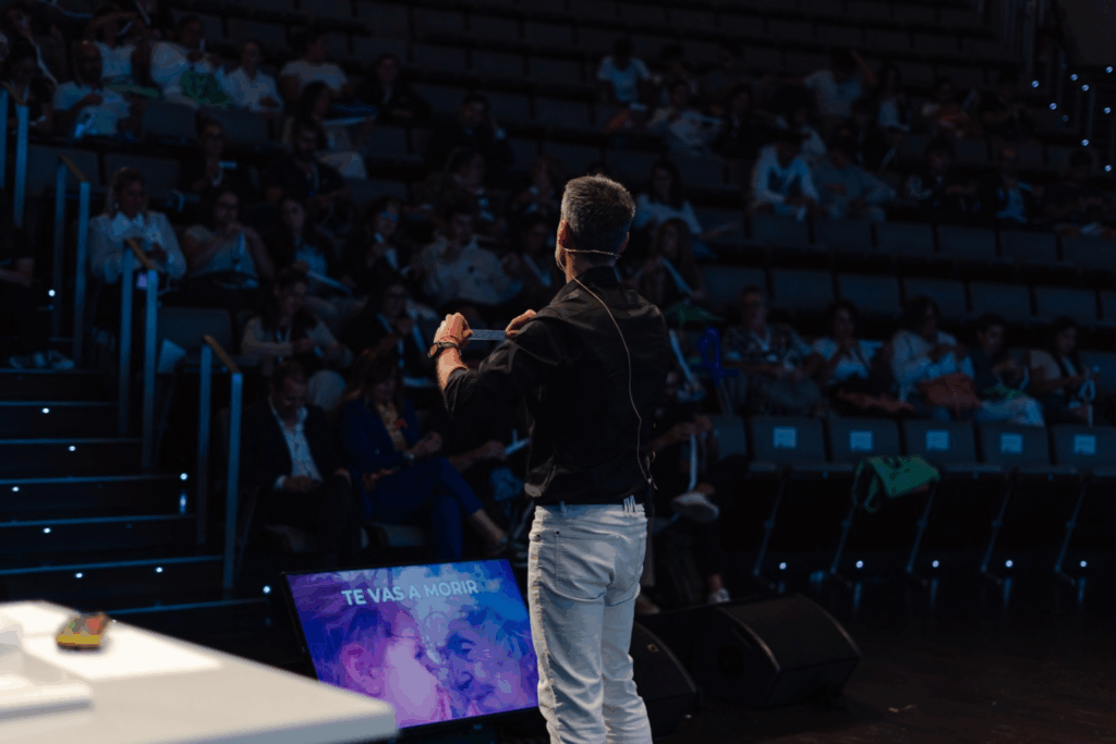 Fotografía de Antoni Fernández i Brugués de espaldas, hablando frente a una audiencia en un auditorio, con una pantalla que muestra el mensaje "Te vas a morir" junto a una imagen emotiva de un niño y una persona mayor.