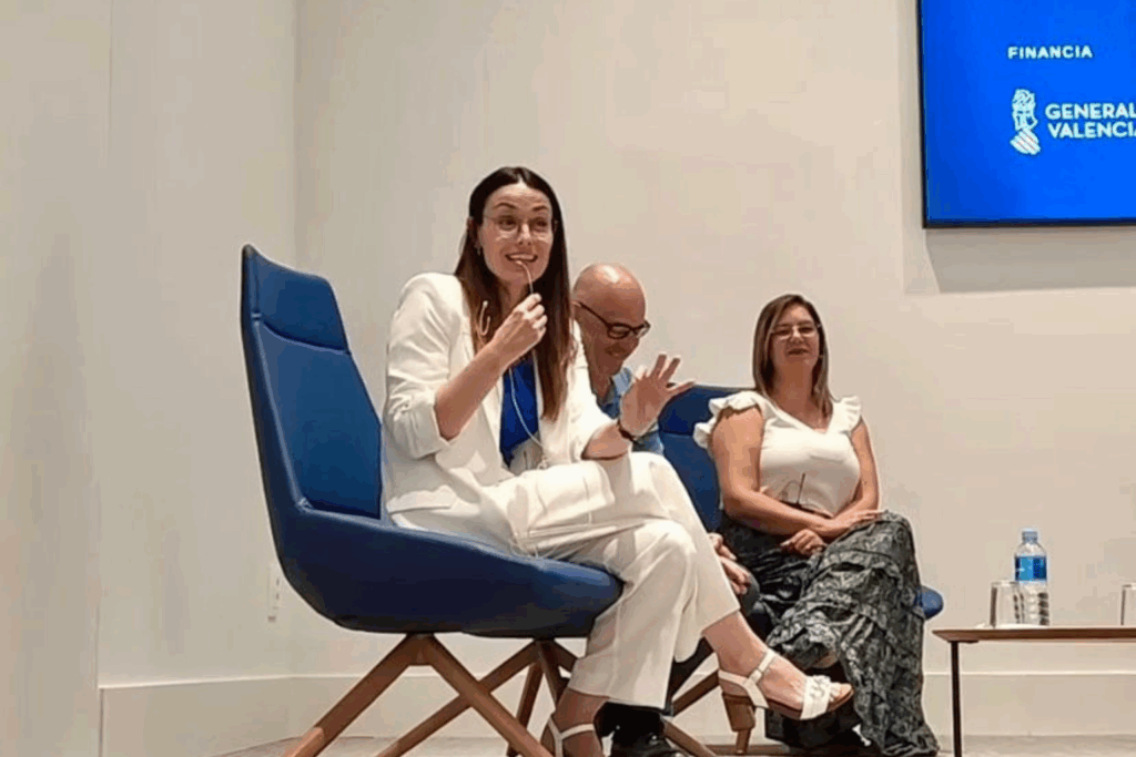Fotografía de Eva Mazuecos durante una conferencia, sentada en un sillón azul con micrófono en mano, vestida de blanco y acompañada de dos colegas, en un espacio moderno y minimalista patrocinado por la Generalitat Valenciana.