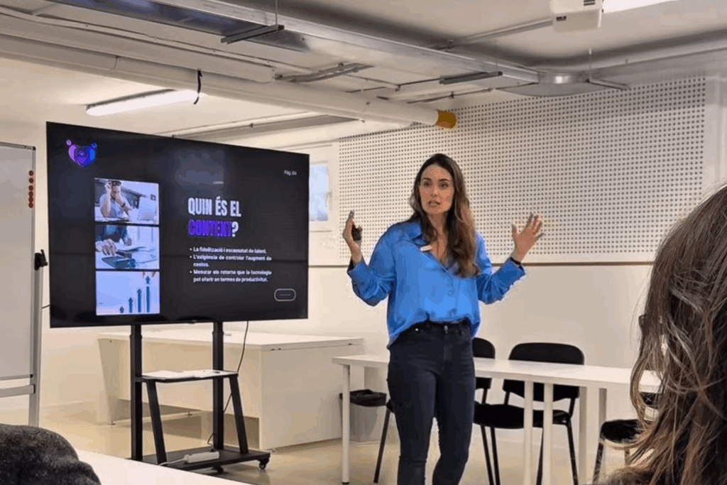 Fotografía de Eva Mazuecos impartiendo una conferencia en un aula moderna, vestida con blusa azul brillante y jeans oscuros, de pie junto a una pantalla con presentación en catalán sobre talento y productividad.