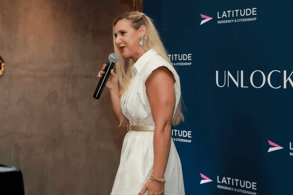 Fotografía de Melinda Alemán durante una conferencia, vestida con un elegante conjunto blanco y hablando con micrófono en mano frente a un panel de fondo corporativo.