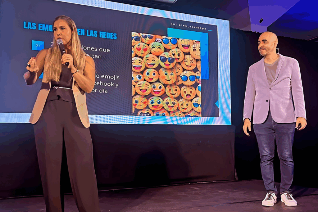 Fotografía de Melinda Alemán presentando en el escenario junto a un conferencista masculino, con una pantalla detrás que muestra emojis y datos sobre redes sociales.