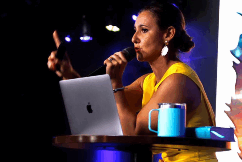 Fotografía de Melinda Alemán hablando con micrófono desde una mesa con laptop, taza y libreta, en un escenario con luces tenues.