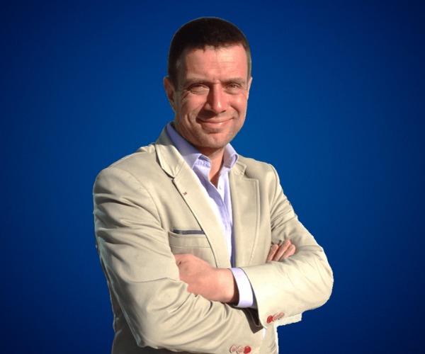 Fotografía de Mauricio Espina, con fondo azul sólido, vistiendo saco beige claro y camisa blanca, posando con los brazos cruzados y una expresión confiada.