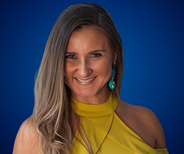 Fotografía de Melinda Alemán con fondo azul, luciendo un vestido amarillo sin mangas y unos pendientes turquesa, sonriente y mirando al frente.