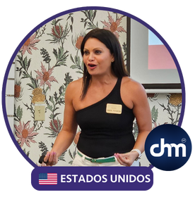 Fotografía de Yanira Puy durante una conferencia en interiores, luciendo una blusa negra de un solo hombro, expresiva y en actitud de liderazgo frente a un proyector y un fondo decorado con papel tapiz floral.