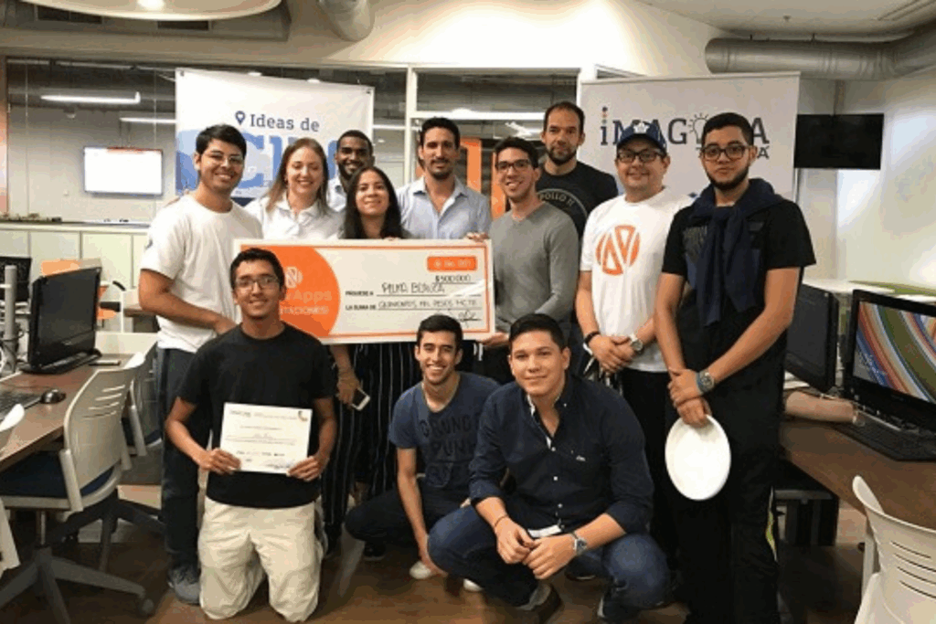 Fotografía de Gustavo De La Vega, conferencista colombiano, posando junto a un grupo de jóvenes emprendedores en un espacio de coworking, sosteniendo un cheque simbólico de premiación en un concurso de innovación.