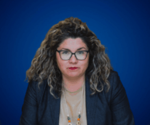 Fotografía de Geraldine Manríquez sobre fondo azul, con chaqueta oscura, blusa beige y collar colorido; expresión seria y profesional, con gafas y cabello rizado.